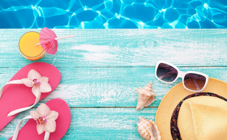 Imagem de piscina com apetrechos de verão próximos, como óculos escuros, chapéu e sandálias.