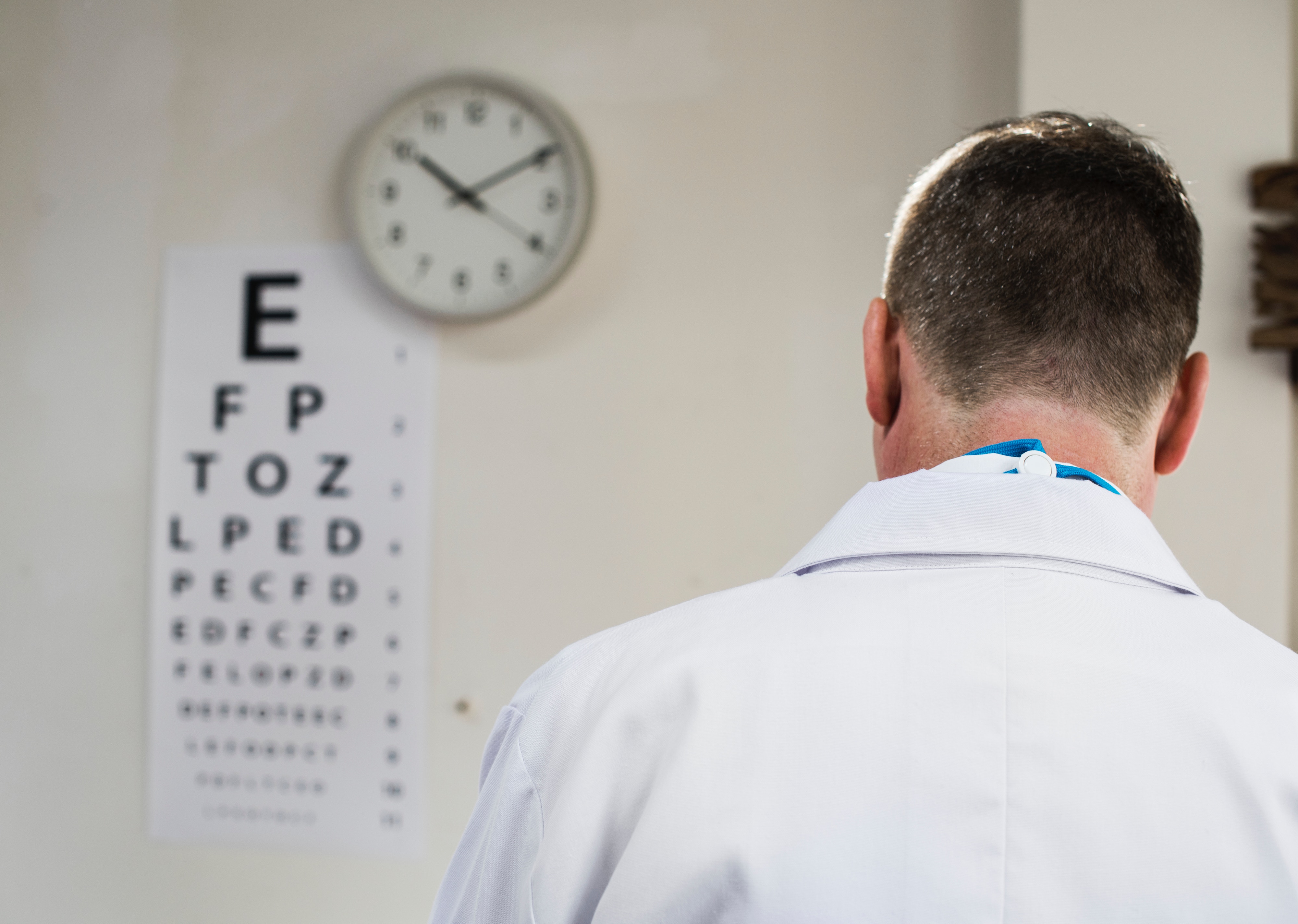 médico de vista olhando para a parede, onde tem um relógio e um cartaz com letras de diferentes tamanhos