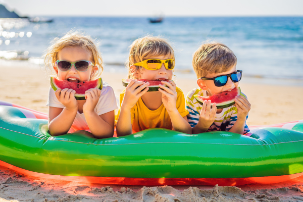 crianças podem usar óculos escuros? na foto, três delas estão na praia, comendo melancia, sorrindo e usando óculos de sol