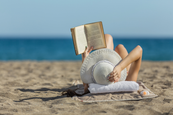 mulher caucasiana de chapéu branco, lendo na areia da praia, ilustra artigo sobre leitura e saúde ocular