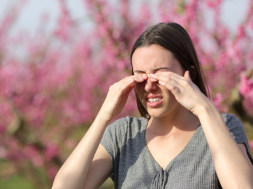 mulher branca em uma paisagem desfocada com flores rosadas atrás, está com as mãos coçando os olhos; demonstra Como se prevenir e cuidar das doenças oculares da primavera.