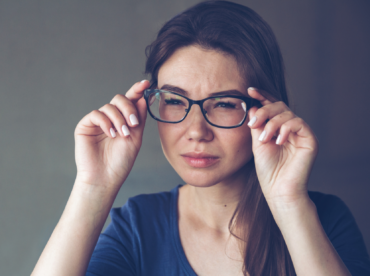 Sobre fundo cinza, uma mulher segura o óculos de hastes pretas que está em seu rosto, olhando adiante, com o cenho franzido; exemplificando os problemas oculares causados pelo estresse.