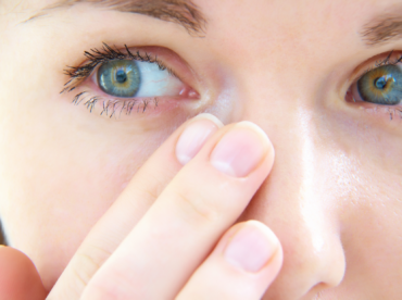 Em close está o rosto de uma mulher branca, de olhos azuis esverdeados e centro amarelado, olhando para o lado e com as pontas dos dedos de uma mão próximas ao canto do olho direito; exemplifica o que pode causar a obstrução do canal lacrimal.