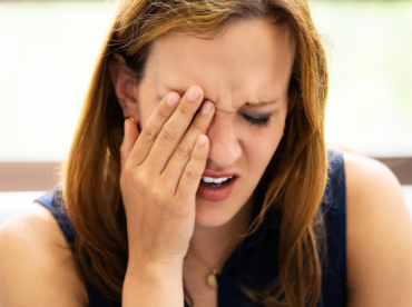 Fotografia de uma mulher branca, loira, aparecendo o rosto em destaque, com uma das mãos sobre o olho direito e o cenho franzido, indicando dor ou incômodo representativo dos sintomas da conjuntivite.