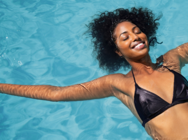 mulher jovem negra, de biquíni preto, de olhos fechados e sorridente, flutua em águas límpidas para ilustrar artigo sobre piscina e vermelhidão nos olhos