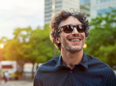 Homem branco sorridente usando óculos escuros em ambiente urbano ilustra artigo sobre consequências dos raios ultravioleta na saúde da visão