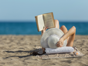 mulher caucasiana de chapéu branco, lendo na areia da praia, ilustra artigo sobre leitura e saúde ocular